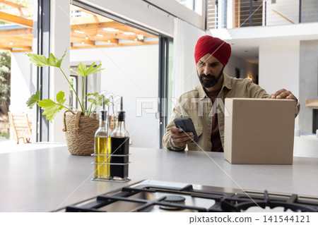 Middle-aged asian indian man sitting at kitchen counter holding smartphone and box next to cooktop Middle-aged asian indian man sitting at kitchen counter holding smartphone and box next to cooktop 141544121