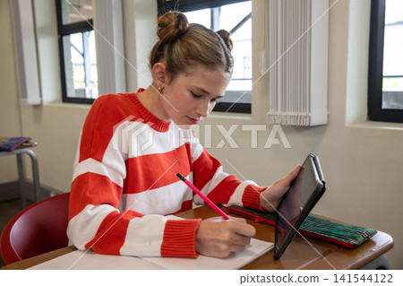 White girl sitting at school desk writing in notebook using pink pencil and holding tablet White girl sitting at school desk writing in notebook using pink pencil and holding tablet 141544122