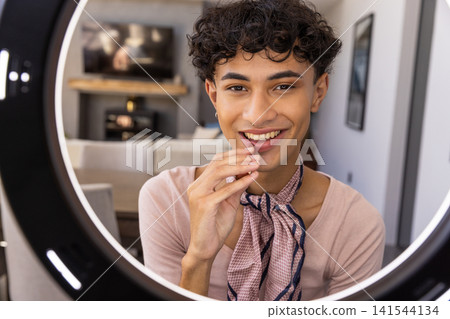Man in mid-twenties wearing scarf smiling into circular led ring light in living room Man in mid-twenties wearing scarf smiling into circular led ring light in living room 141544134