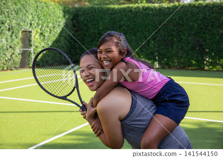 Asian mom carrying daughter piggyback on turf tennis court with boundary lines while holding racket 141544150