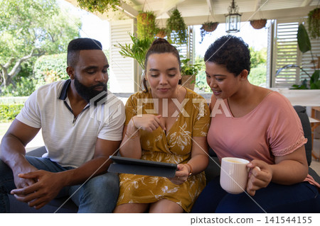 Diverse friends sitting on sofa under white pergola while pointing at tablet and holding coffee mug Diverse friends sitting on sofa under white pergola while pointing at tablet and holding coffee mug 141544155