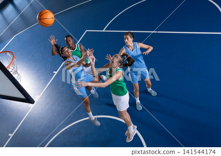 Diverse female opponents jumping and reaching for orange basketball under hoop on blue court Diverse female opponents jumping and reaching for orange basketball under hoop on blue court 141544174