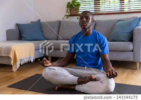 African american man meditating on black yoga mat in living room with sofa, blue pillows African american man meditating on black yoga mat in living room with sofa, blue pillows 141544175