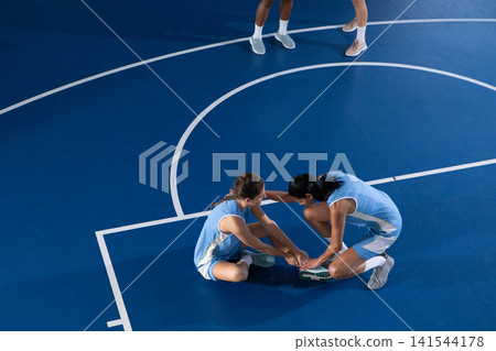 Diverse female basketball teammates stretching forward while seated on blue court wearing uniforms 141544178