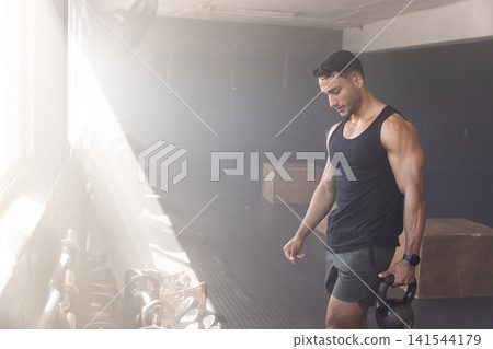 Male athlete lifting kettlebell in gym studio with kettlebell rack and plyometric boxes, copy space 141544179