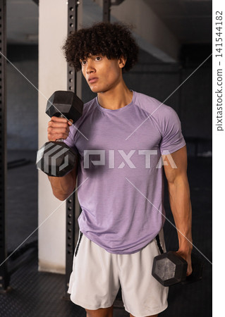 Man lifting hexagonal dumbbells at waist height in gym workout area near weight rack Man lifting hexagonal dumbbells at waist height in gym workout area near weight rack 141544182