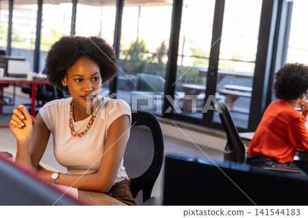 Diverse female coworkers sitting on ergonomic chairs at desk in office holding pen by monitors Diverse female coworkers sitting on ergonomic chairs at desk in office holding pen by monitors 141544183