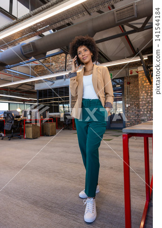 African american woman walking through office talking on smartphone next to desks African american woman walking through office talking on smartphone next to desks 141544184