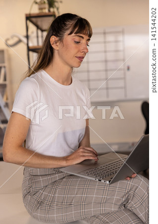 Mid adult woman leaning forward working on laptop at office desk with whiteboard, shelves, plant Mid adult woman leaning forward working on laptop at office desk with whiteboard, shelves, plant 141544202