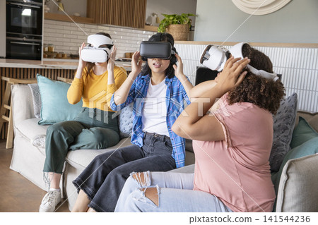 Diverse female friends adjusting VR headsets sitting on couch with cushions next to kitchen Diverse female friends adjusting VR headsets sitting on couch with cushions next to kitchen 141544236