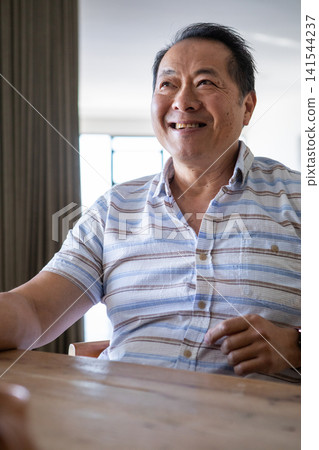 Senior asian man wearing shirt smiling while sitting at home dining table by window light 141544237