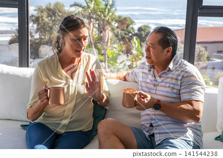 Diverse senior couple holding ceramic mugs and chatting on sofa in coastal living room Diverse senior couple holding ceramic mugs and chatting on sofa in coastal living room 141544238