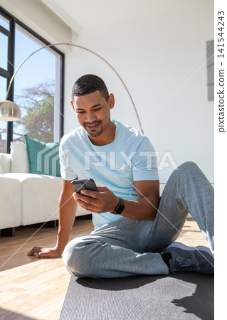 Man in his thirties sitting cross-legged on black exercise mat at home holding smartphone Man in his thirties sitting cross-legged on black exercise mat at home holding smartphone 141544243