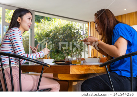 Asian mother and daughter eating from bowls with chopsticks at wooden dining table in living room Asian mother and daughter eating from bowls with chopsticks at wooden dining table in living room 141544256
