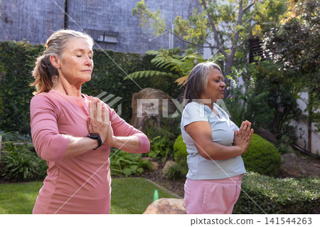 Diverse senior women friends meditating in garden wearing pink top and shirt with black watch Diverse senior women friends meditating in garden wearing pink top and shirt with black watch 141544263