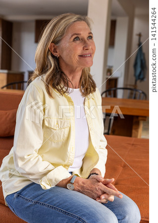 Senior woman sitting on sofa cushion in living room gazing upward showing wristwatch, bracelet Senior woman sitting on sofa cushion in living room gazing upward showing wristwatch, bracelet 141544264