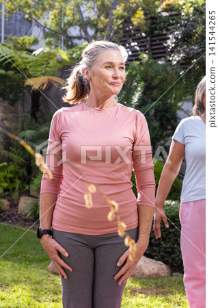 Senior female friends exercising in backyard wearing pink tops and smartwatch Senior female friends exercising in backyard wearing pink tops and smartwatch 141544265