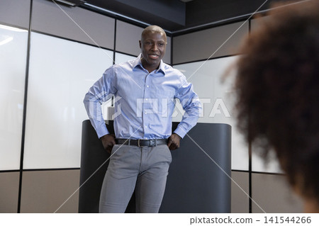 Diverse coworkers standing at podium speaking toward colleague in office with glass panels Diverse coworkers standing at podium speaking toward colleague in office with glass panels 141544266
