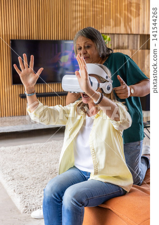 Diverse senior female friends trying out virtual reality headset on bench in living room Diverse senior female friends trying out virtual reality headset on bench in living room 141544268
