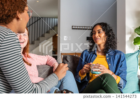 Diverse female friends sitting on couch in living room holding ceramic mug and throw pillows 141544273
