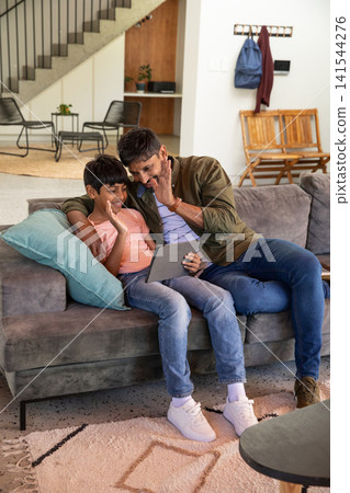 South asian father and son waving at tablet while sitting on sofa in living room South asian father and son waving at tablet while sitting on sofa in living room 141544276
