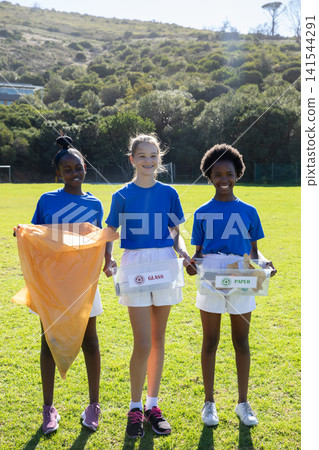 Diverse teen girls standing on sports field holding trash bag with glass and paper bins Diverse teen girls standing on sports field holding trash bag with glass and paper bins 141544291