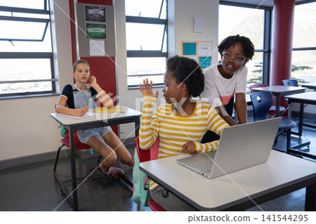 Diverse children sitting at desks in classroom raising hand by laptop writing in notebook Diverse children sitting at desks in classroom raising hand by laptop writing in notebook 141544295