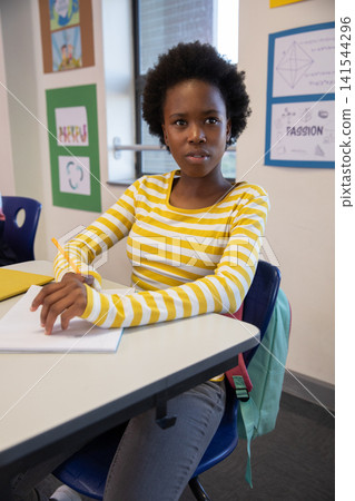 African american girl student writing in notebook at school desk with pencil, backpack, posters African american girl student writing in notebook at school desk with pencil, backpack, posters 141544296