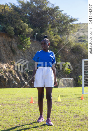 African american female athlete wearing sportswear warming up on soccer pitch with cones, goal 141544297