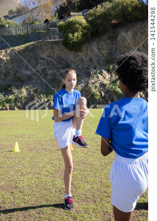 Diverse teenage teammates stretching on green sports field near cone wearing sneakers Diverse teenage teammates stretching on green sports field near cone wearing sneakers 141544298