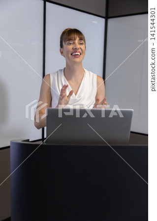 Woman presenting talk at conference room using laptop, lectern with whiteboard background Woman presenting talk at conference room using laptop, lectern with whiteboard background 141544301