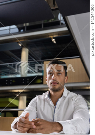 Businessman sitting at wooden table in office with columns, glass railing and acoustic panels Businessman sitting at wooden table in office with columns, glass railing and acoustic panels 141544306