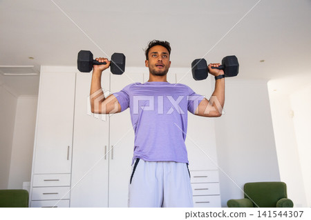 Asian man lifting black hex dumbbells at shoulder height in home gym in purple sportswear Asian man lifting black hex dumbbells at shoulder height in home gym in purple sportswear 141544307
