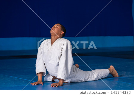 Male martial arts student performing backbend stretch in dojo on blue mat in white gi belt Male martial arts student performing backbend stretch in dojo on blue mat in white gi belt 141544383