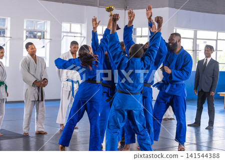 Diverse judo teammates wearing blue gis lifting gold trophy in martial arts hall on mat Diverse judo teammates wearing blue gis lifting gold trophy in martial arts hall on mat 141544388