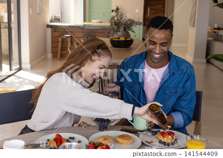 Diverse couple pouring syrup onto pancakes at dining table at home with coffee press 141544409
