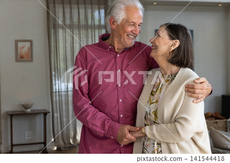 Senior couple hugging and smiling in cozy living room near sheer curtains and woven storage basket Senior couple hugging and smiling in cozy living room near sheer curtains and woven storage basket 141544418