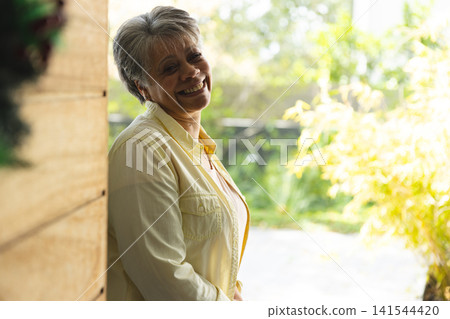 Senior woman leaning against wooden porch wall with wreath facing garden plants smiling in sunlight Senior woman leaning against wooden porch wall with wreath facing garden plants smiling in sunlight 141544420