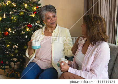 Diverse female friends chatting on sofa by decorated Christmas tree holding ceramic mugs Diverse female friends chatting on sofa by decorated Christmas tree holding ceramic mugs 141544424