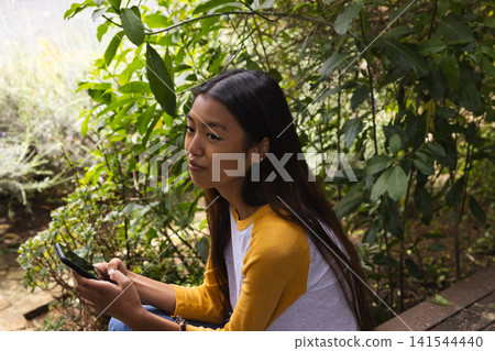 Asian woman sitting on garden wall holding smartphone in lush garden wearing raglan shirt and jeans 141544440