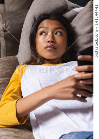 Asian teenage female reclining on brown leather couch in living room holding dark smartphone Asian teenage female reclining on brown leather couch in living room holding dark smartphone 141544441