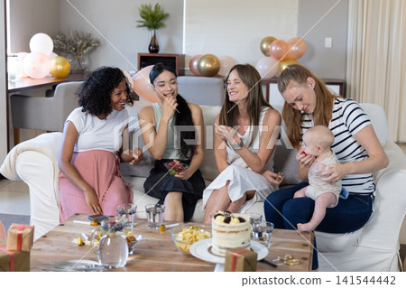 Diverse female friends sitting on sofa in living room sharing layered cake at baby shower 141544442