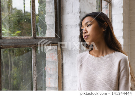 Asian woman standing by paned window with raindrops in white brick loft gazing outside, copy space 141544444