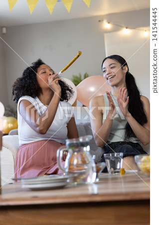 Diverse female friends chatting on sofa in living room, pastel balloons, bunting, water pitcher Diverse female friends chatting on sofa in living room, pastel balloons, bunting, water pitcher 141544445