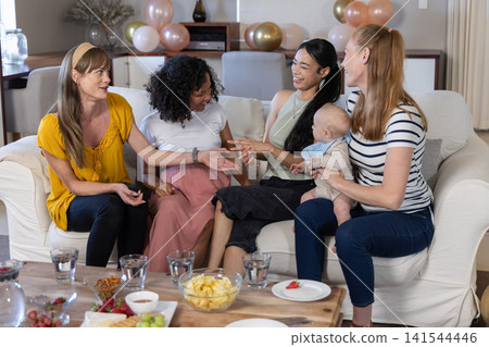 Diverse female friends eating snacks on coffee table while sitting on living room sofa, infant 141544446