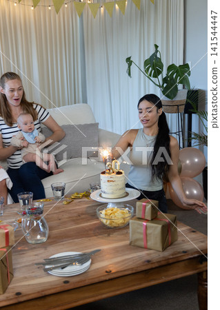 Diverse female friends holding baby while celebrating 30th birthday in living room, cake Diverse female friends holding baby while celebrating 30th birthday in living room, cake 141544447