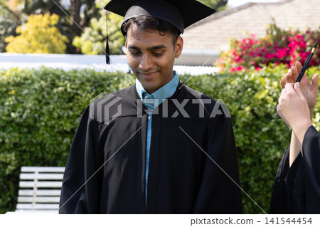 Asian man in graduation gown, mortarboard, examining pen being offered by classmate in backyard Asian man in graduation gown, mortarboard, examining pen being offered by classmate in backyard 141544454