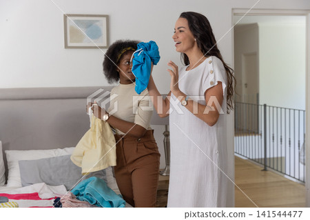 Diverse female friends sorting and folding pile of colorful clothes on bed by nightstand lamp Diverse female friends sorting and folding pile of colorful clothes on bed by nightstand lamp 141544477