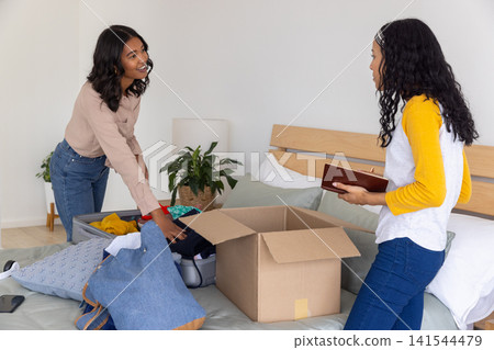 Diverse mom and teenage daughter standing by bed packing clothes from open suitcase into box 141544479