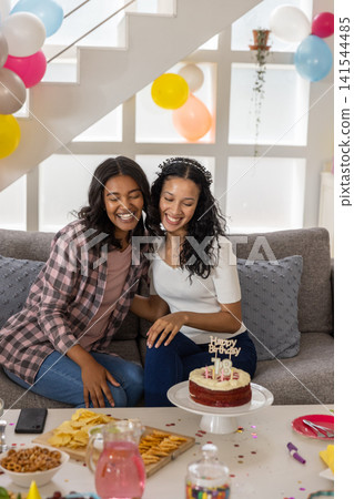 Diverse mom and teenage daughter sitting on sofa by stairs celebrating birthday with cake balloons Diverse mom and teenage daughter sitting on sofa by stairs celebrating birthday with cake balloons 141544485